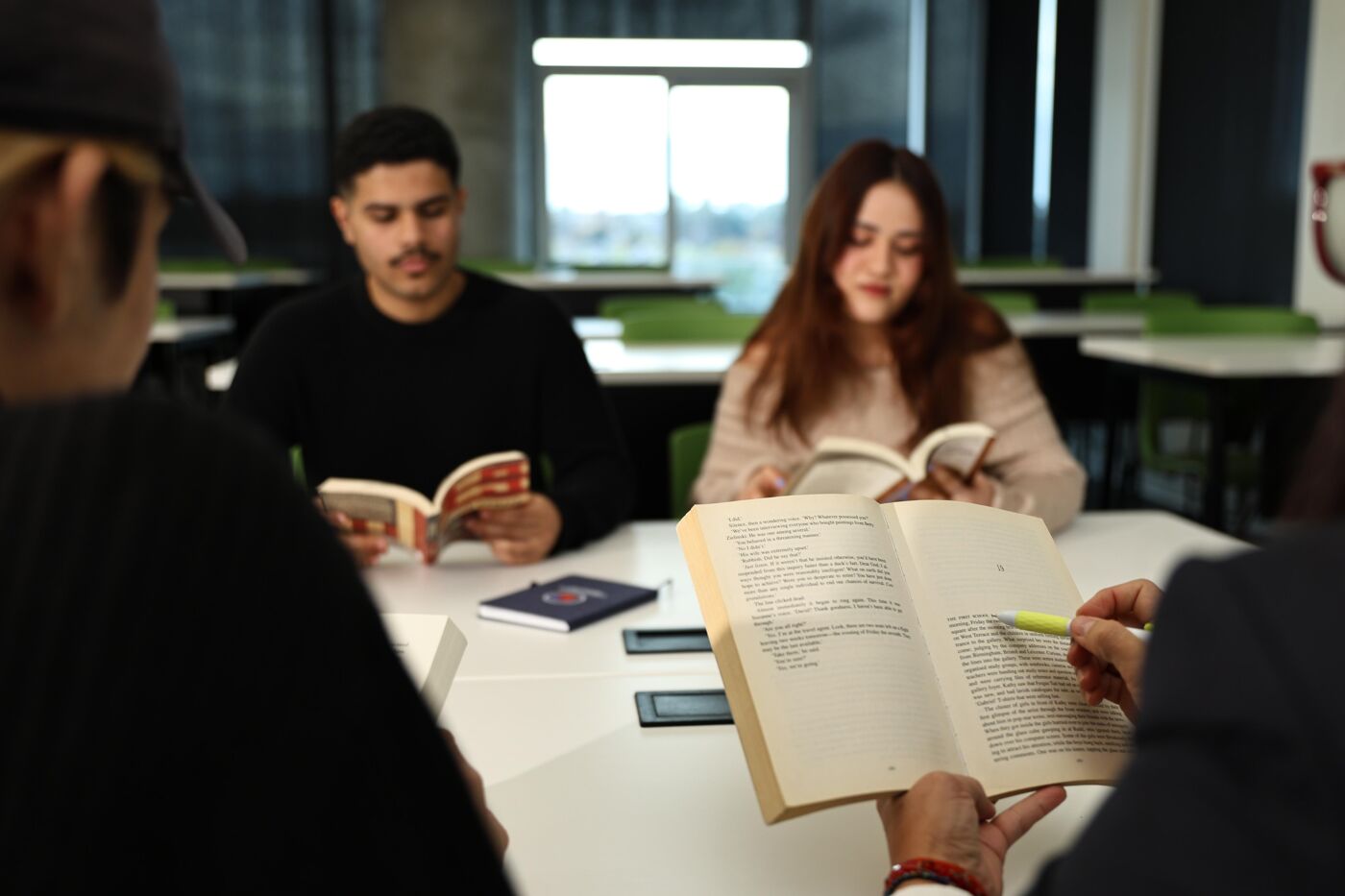 The image shows a group of students sitting around a table, deeply engrossed in reading books. The atmosphere suggests a focused study session, with each individual concentrating on their respective texts.