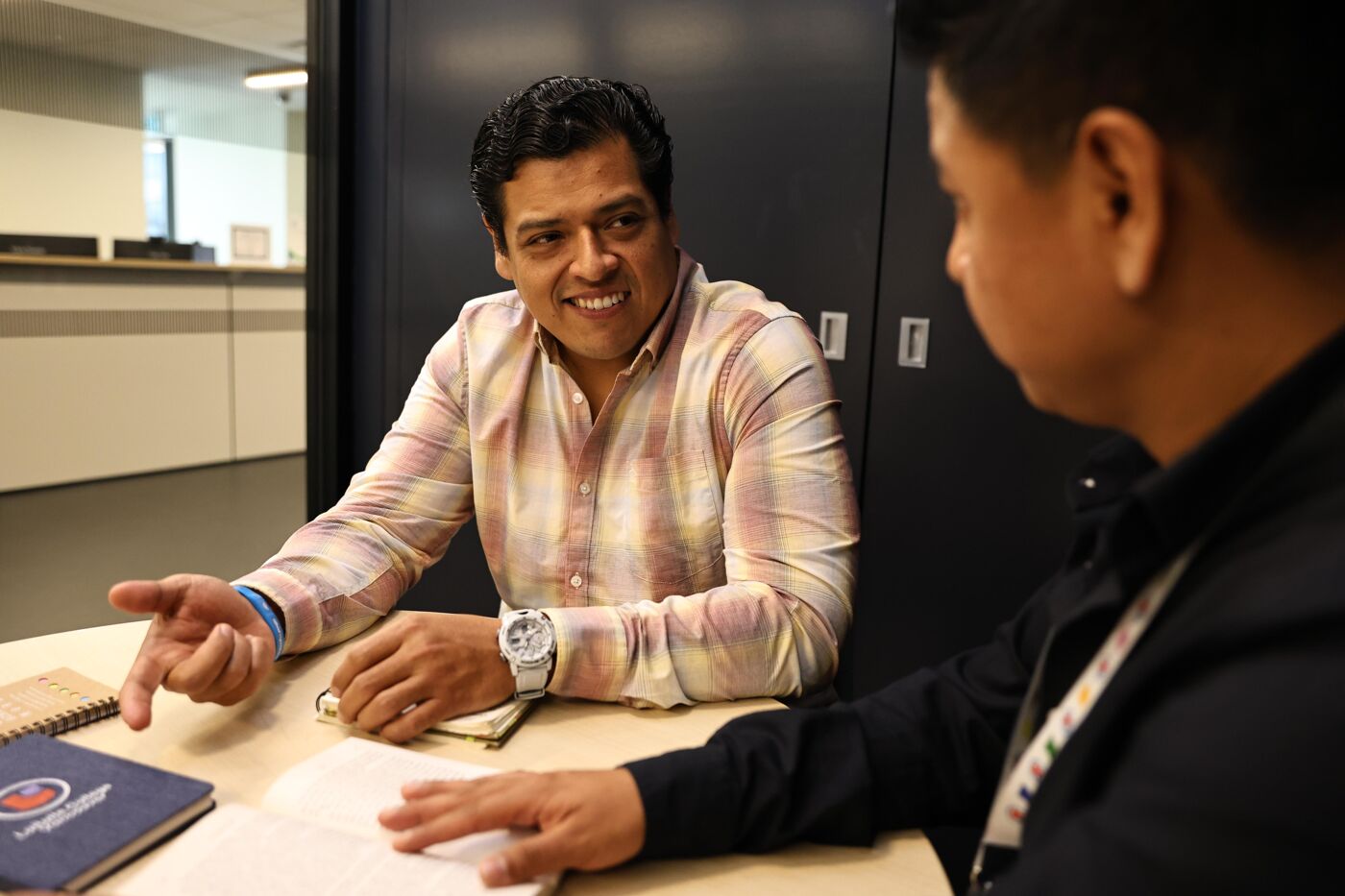 The image shows two men in an office setting, seemingly engaged in a discussion. One man is wearing a patterned shirt and a watch, gesturing with his hand, while the other man is partially visible, listening attentively. There are notebooks on the table.