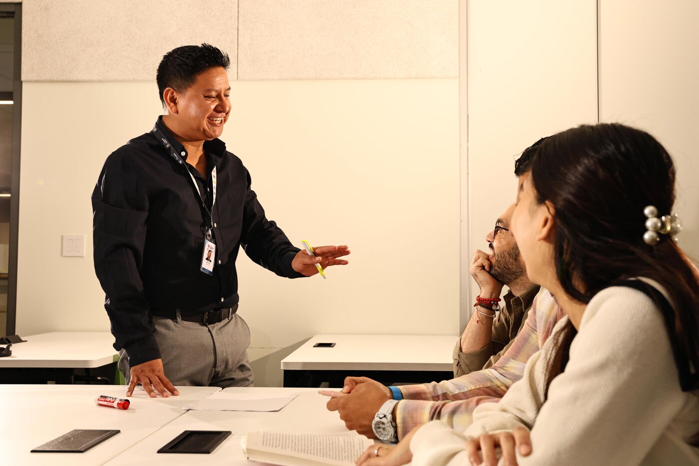 A man in a black shirt is standing and gesturing with his right hand while addressing two people seated at a table. The setting appears to be an office or meeting room.