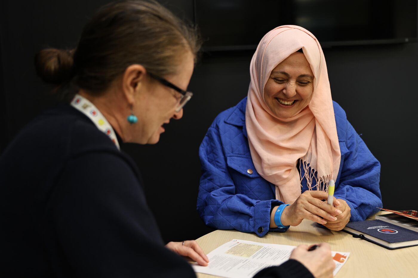 The image shows two women sitting at a table, seemingly engaged in a discussion or meeting. One woman is wearing a hijab and a blue jacket, while the other is wearing glasses and a dark top. They both appear to be looking at a book or document on the table.