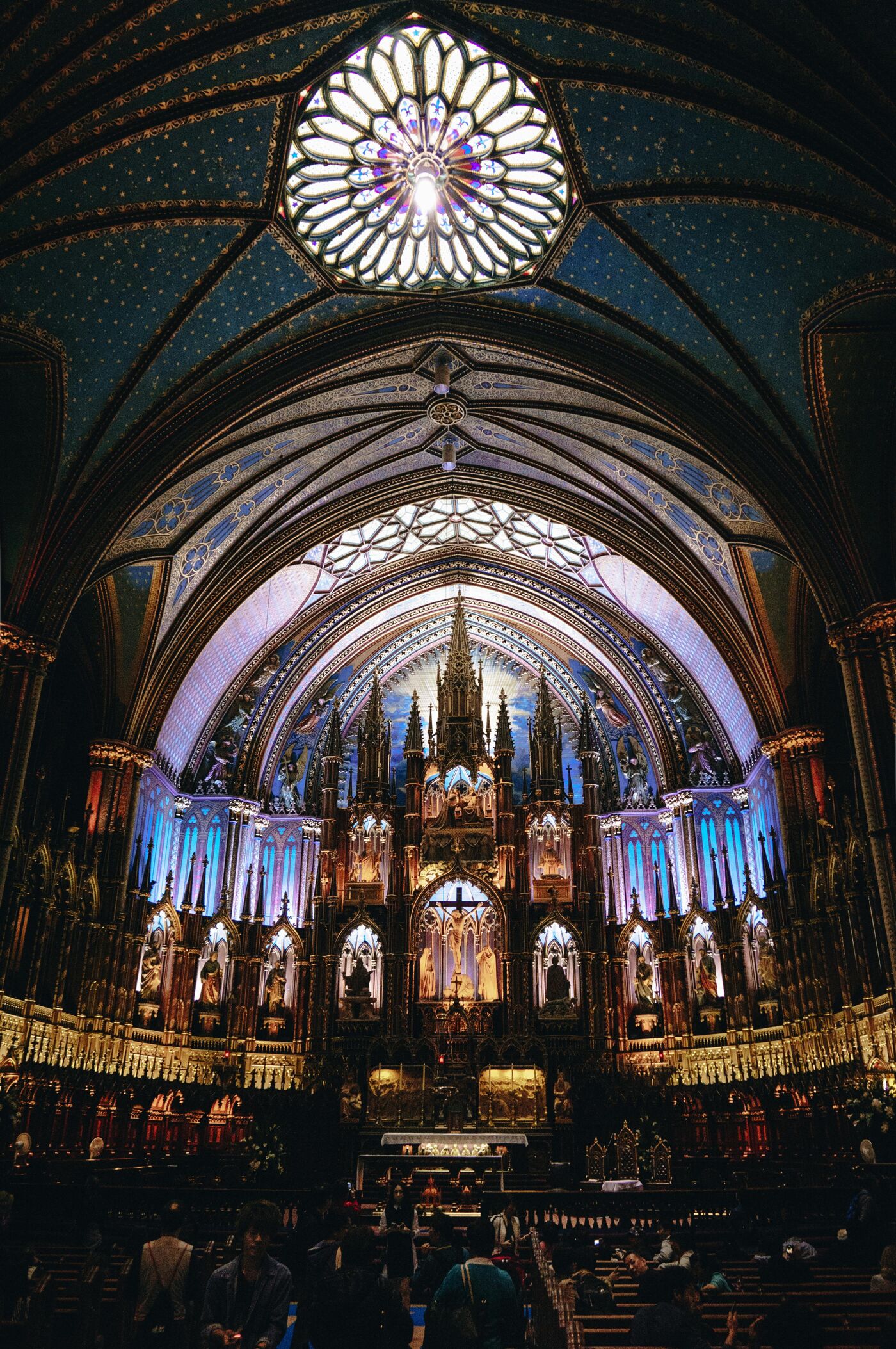 The image shows the interior of a grand cathedral, featuring ornate Gothic architecture, including pointed arches, ribbed vaults, and a large rose window. The space is illuminated by a combination of natural and artificial light, highlighting the intricate details of the building's design.
