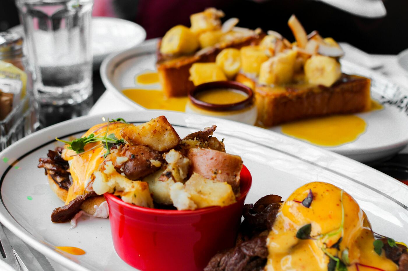 The image shows a close-up of two plates of food. One plate has what appears to be french toast topped with bananas and sauce. The other plate features a dish with potatoes, meat, and an egg with hollandaise sauce.