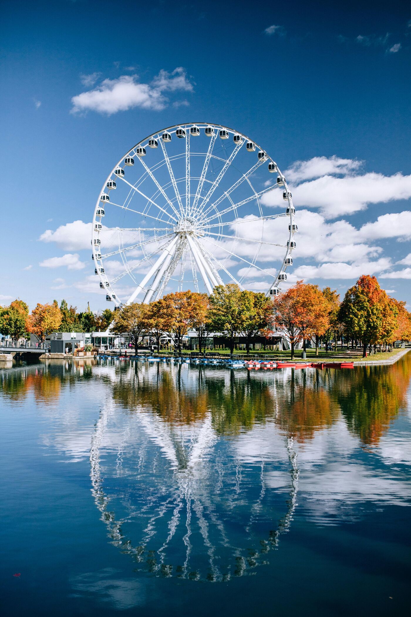 The image shows a large Ferris wheel reflected in calm water. Trees with autumn foliage line the waterfront, and a blue sky with scattered clouds is visible in the background. The scene is peaceful and picturesque.
