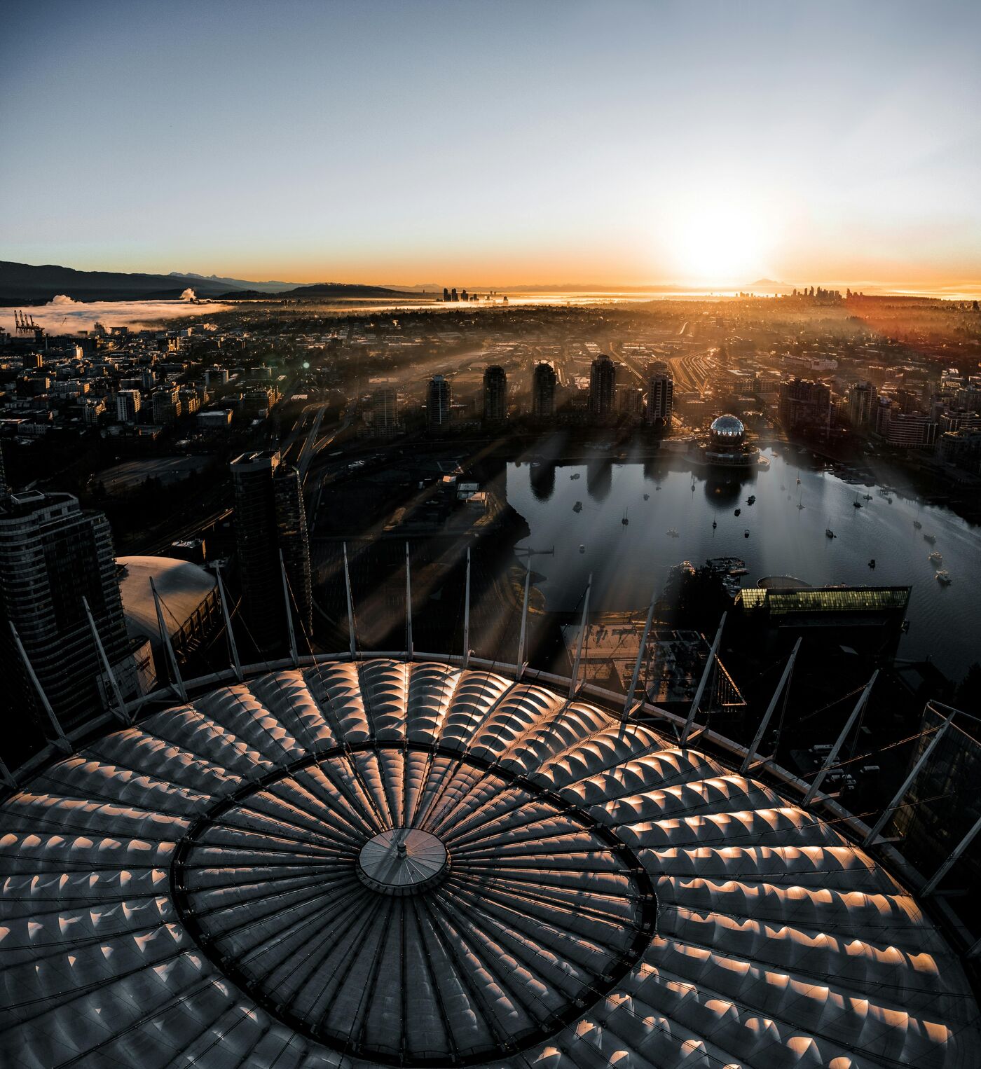 The image shows a cityscape at sunset, viewed from a high vantage point, possibly a rooftop. The sun is setting over the horizon, casting a warm glow over the city and the water. The foreground features a circular structure with a ribbed pattern.