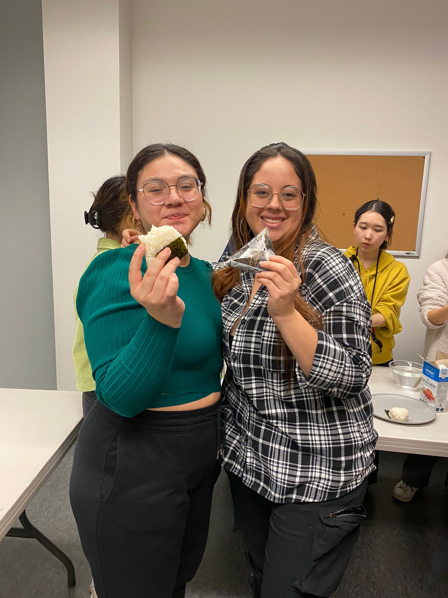 Two women are smiling and holding what appears to be onigiri or rice balls. They are standing indoors, possibly at a food-related event or cooking class, with other people in the background.