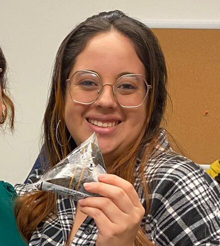 Two women are smiling and holding what appears to be onigiri or rice balls. They are standing indoors, possibly at a food-related event or cooking class, with other people in the background.