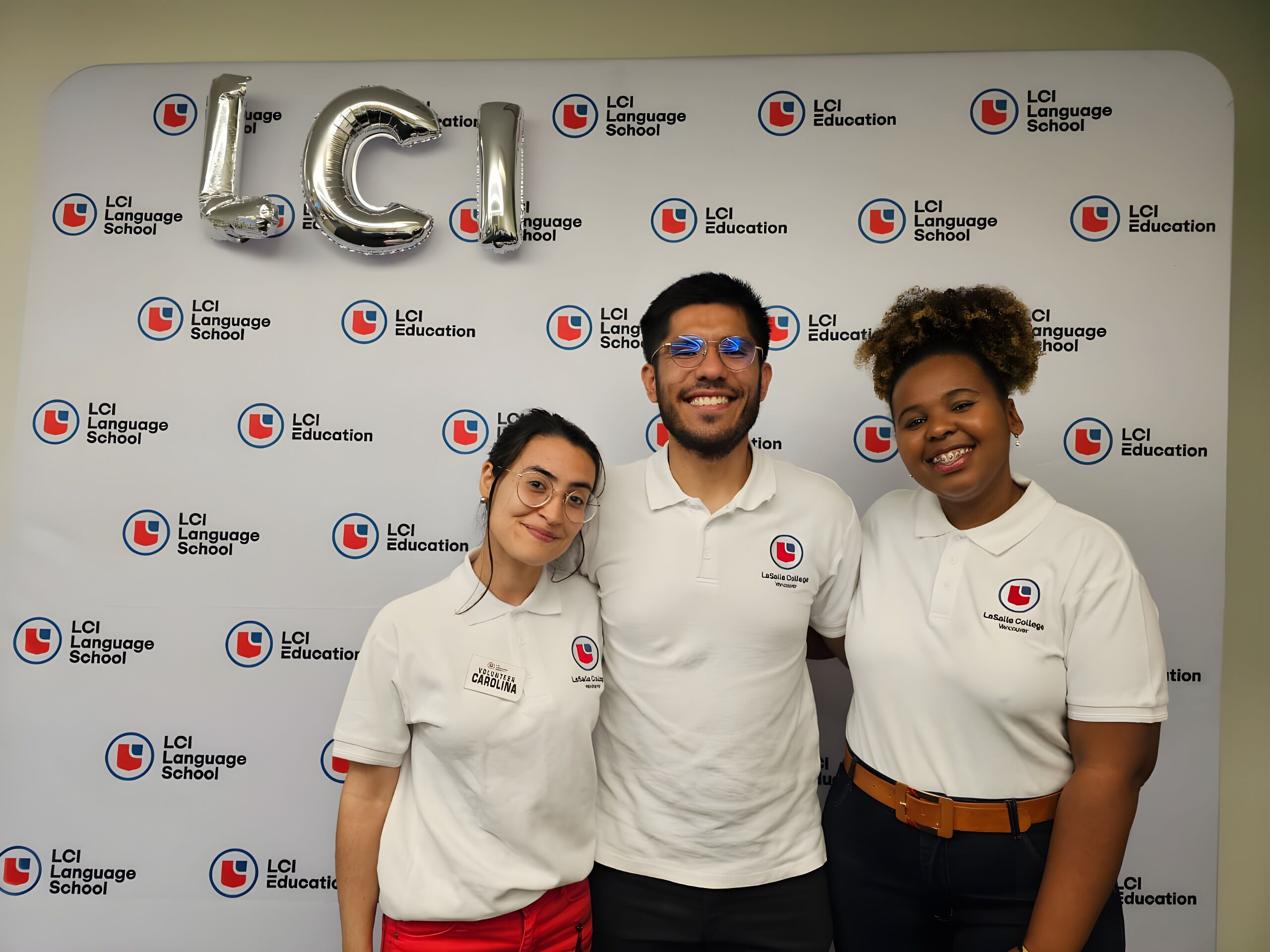Three individuals are posing in front of a backdrop adorned with the logo of LCI Language School. The backdrop also features silver balloon letters spelling out &quot;LCI&quot;. The people in the image are smiling and appear to be associated with the language school.