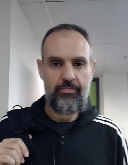 The image shows a man with a beard and short dark hair, wearing a black hoodie with white stripes on the sleeves. He appears to be in a hallway with a backpack slung over his shoulder.