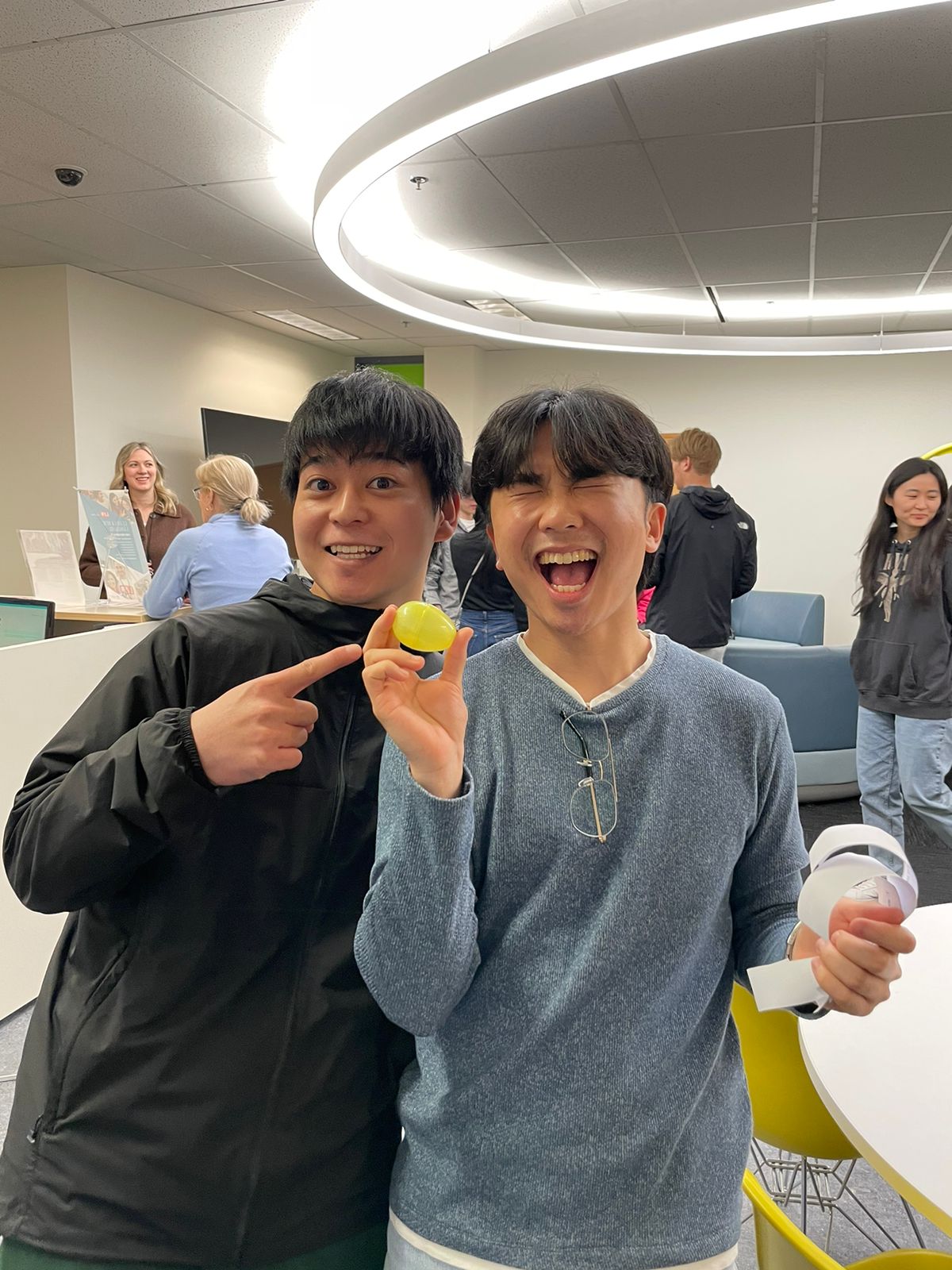 Two smiling men pose indoors, one pointing at a yellow ball held by the other. They are in a brightly lit room with other people and furniture visible in the background.