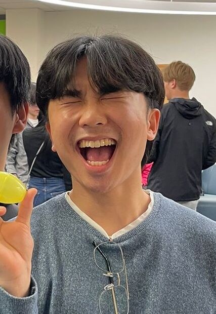 Two smiling men pose indoors, one pointing at a yellow ball held by the other. They are in a brightly lit room with other people and furniture visible in the background.