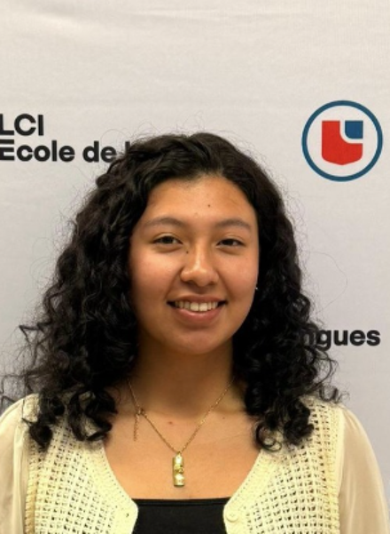 A young woman with curly hair stands in front of a backdrop featuring the LCI Education logo. She is wearing a black top and a light-colored, patterned cardigan. She has a necklace and bracelets on. She is smiling and looking at the camera.