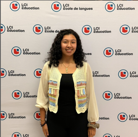 A young woman with curly hair stands in front of a backdrop featuring the LCI Education logo. She is wearing a black top and a light-colored, patterned cardigan. She has a necklace and bracelets on. She is smiling and looking at the camera.