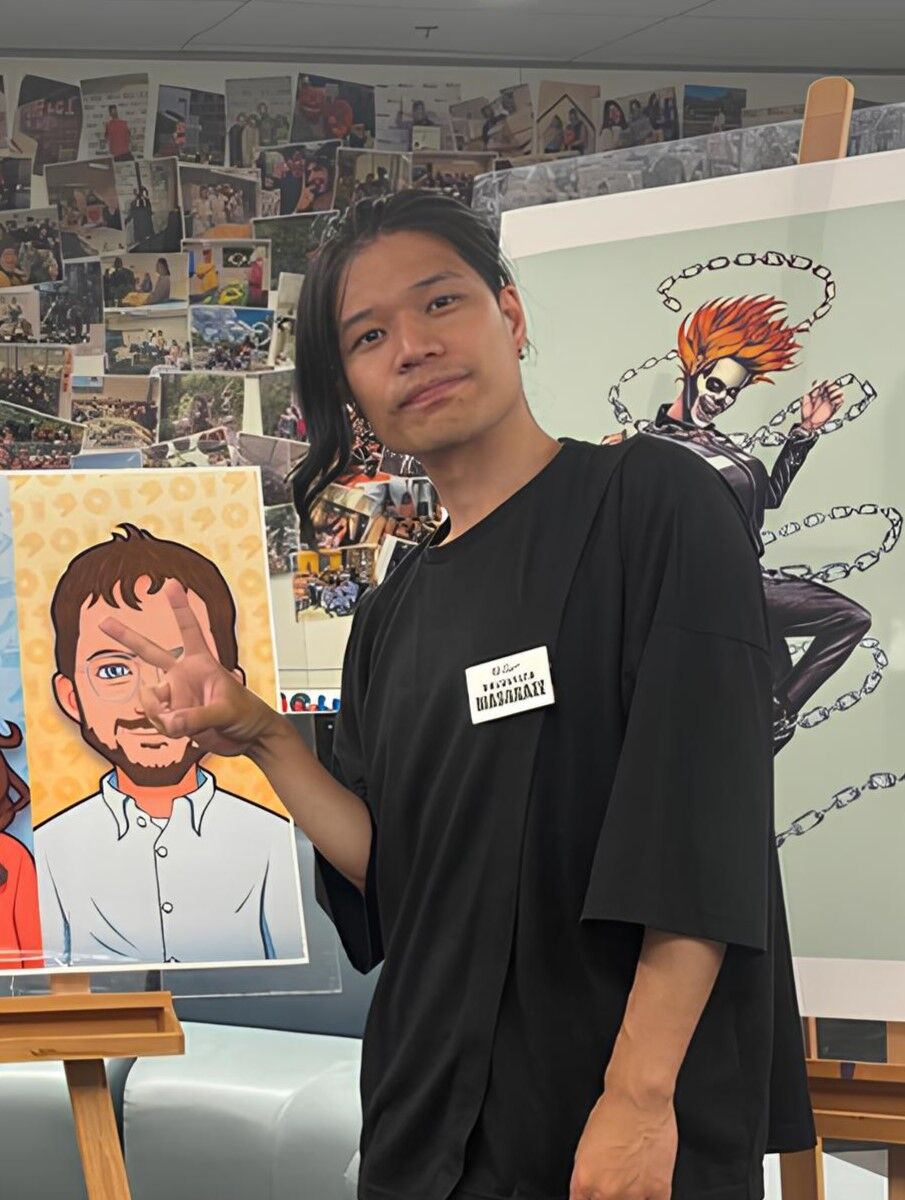 A man with long dark hair is standing in front of two cartoon drawings. He is wearing a black shirt and holding up three fingers. There are many pictures on the wall behind him.