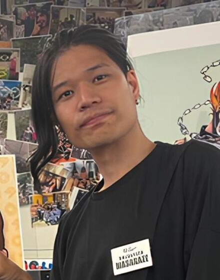 A man with long dark hair is standing in front of two cartoon drawings. He is wearing a black shirt and holding up three fingers. There are many pictures on the wall behind him.