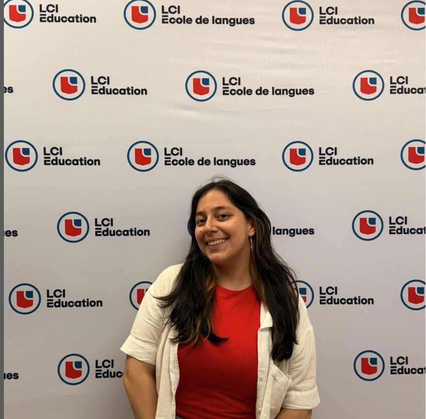 A woman stands smiling in front of a backdrop featuring the logos of LCI Education and LCI Ecole de langues. She is wearing a red shirt and a light-colored jacket, with dark hair that has some lighter streaks.
