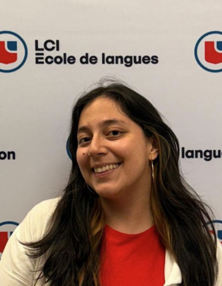 A woman stands smiling in front of a backdrop featuring the logos of LCI Education and LCI Ecole de langues. She is wearing a red shirt and a light-colored jacket, with dark hair that has some lighter streaks.