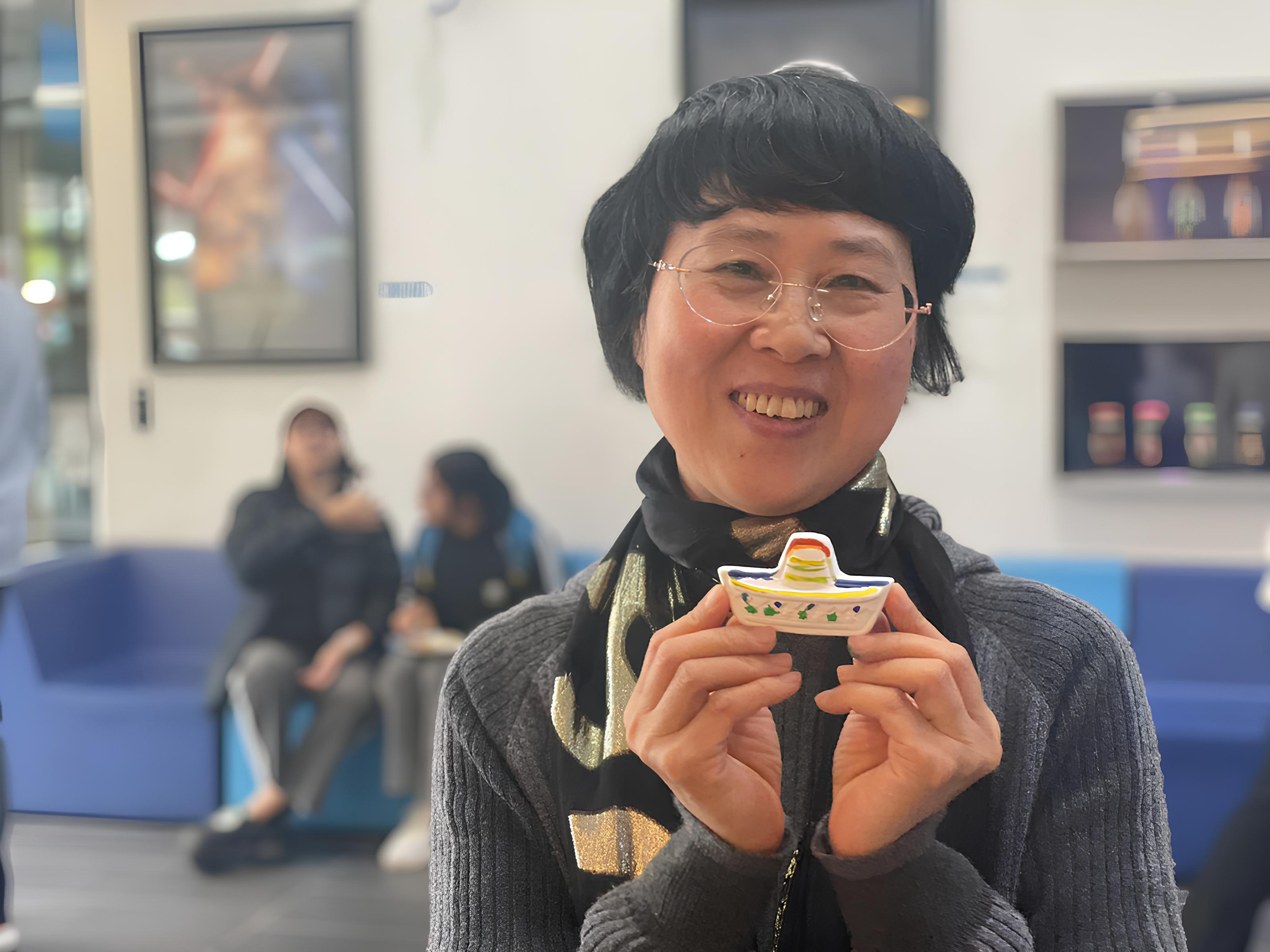 A smiling woman with glasses holds up a small, decorative item. She is wearing a grey sweater and a patterned scarf. In the background, two people are seated on a blue couch in what appears to be a public space.
