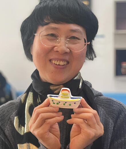 A smiling woman with glasses holds up a small, decorative item. She is wearing a grey sweater and a patterned scarf. In the background, two people are seated on a blue couch in what appears to be a public space.