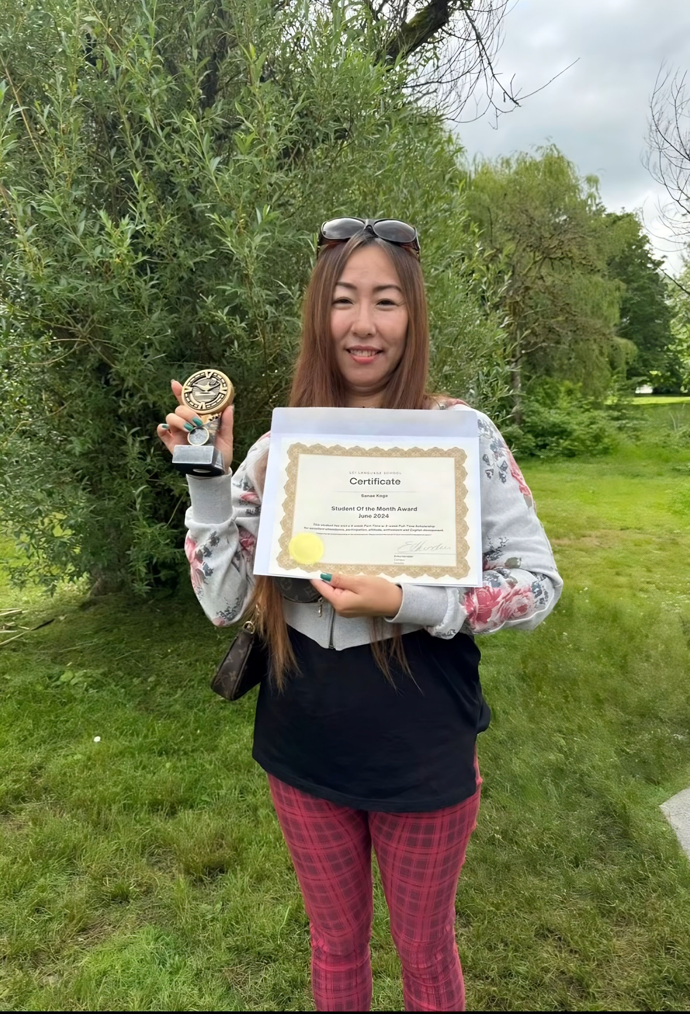 A woman stands outdoors on a grassy lawn, holding a certificate in one hand and a small trophy in the other. She is smiling and wearing sunglasses on her head, with trees and greenery in the background.
