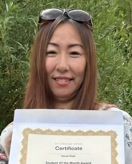 A woman stands outdoors on a grassy lawn, holding a certificate in one hand and a small trophy in the other. She is smiling and wearing sunglasses on her head, with trees and greenery in the background.