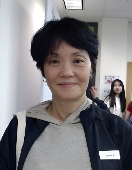 The image shows a middle-aged Asian woman with short black hair, wearing a gray hoodie and a black jacket. She has a name tag that reads "Salina W". There is a young Asian girl in the background.