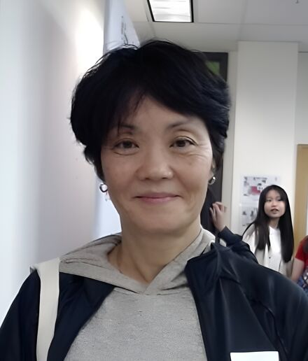 The image shows a middle-aged Asian woman with short black hair, wearing a gray hoodie and a black jacket. She has a name tag that reads "Salina W". There is a young Asian girl in the background.