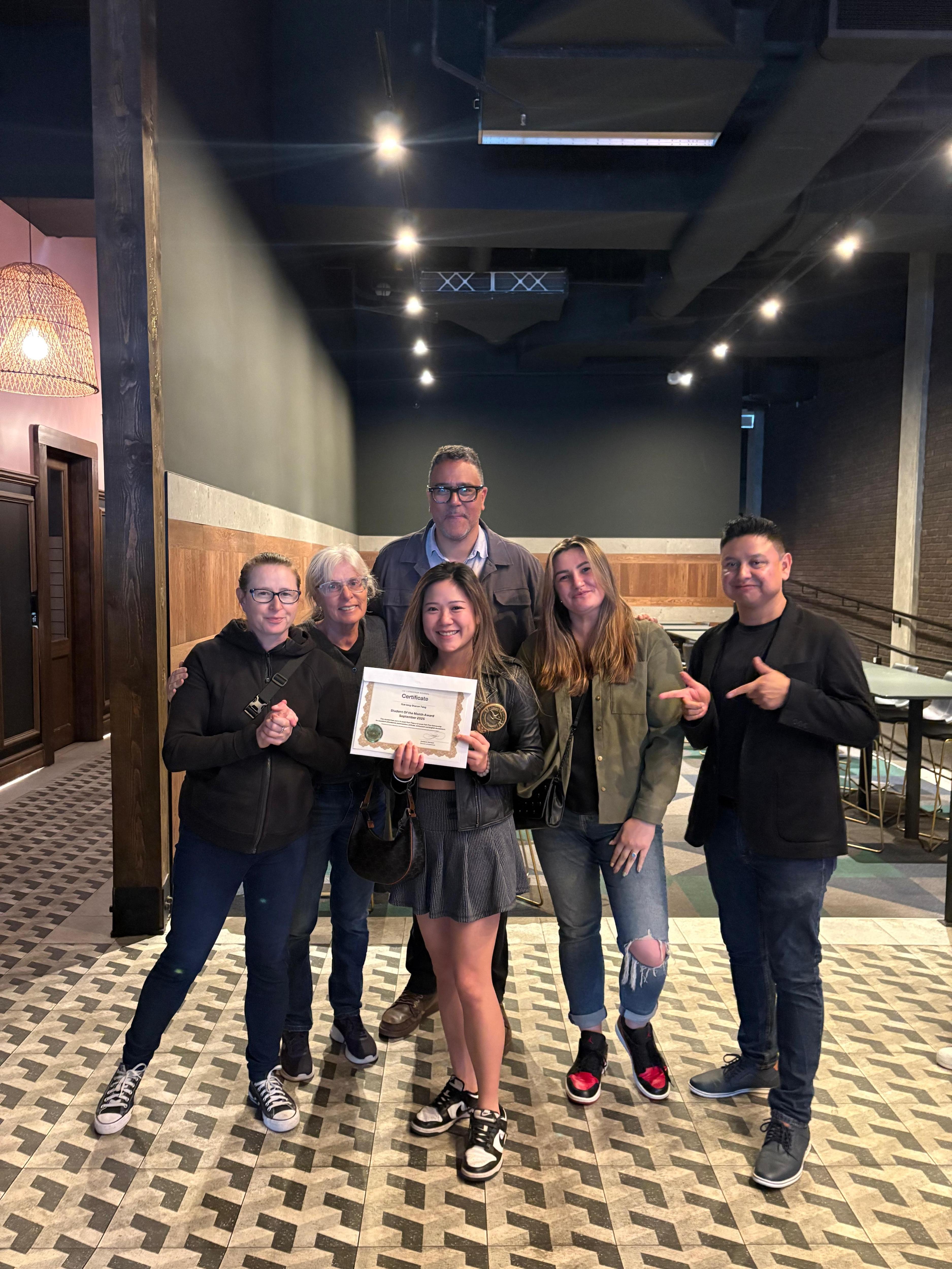 A group of six people are posing for a photo indoors. One person is holding a certificate. They are standing on a patterned floor with a dark background. The lighting is bright, and the atmosphere appears celebratory.