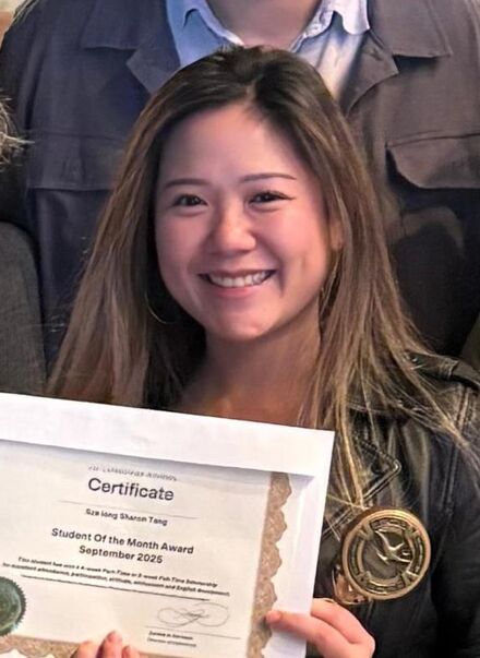 A group of six people are posing for a photo indoors. One person is holding a certificate. They are standing on a patterned floor with a dark background. The lighting is bright, and the atmosphere appears celebratory.