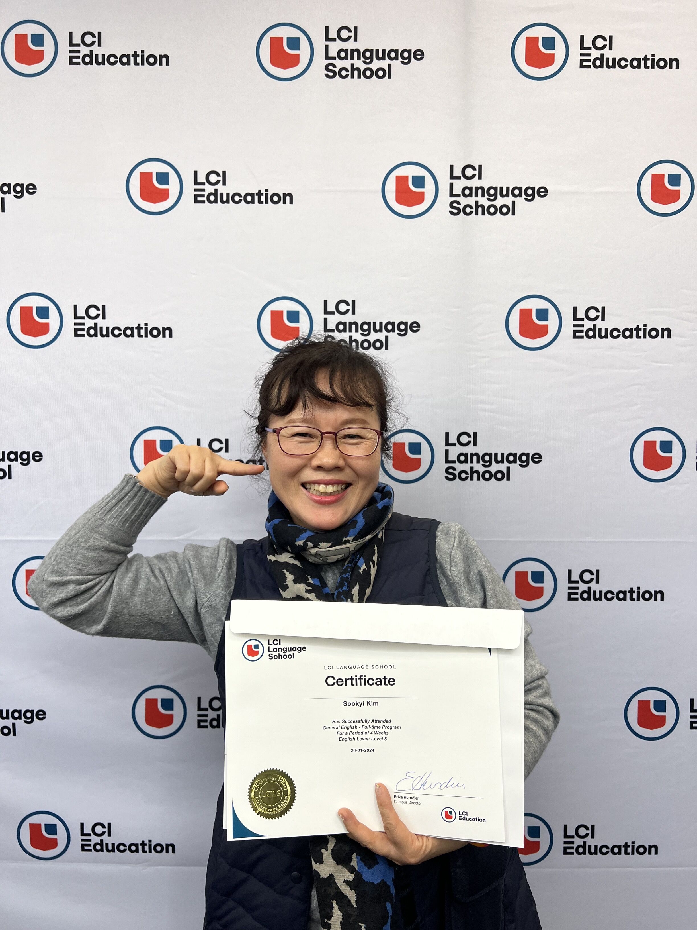 A woman with glasses is holding a certificate in front of an LCI Language School backdrop. She is smiling and pointing to her cheek. The certificate has the LCI Language School logo on it.