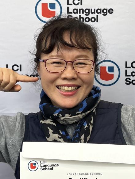 A woman with glasses is holding a certificate in front of an LCI Language School backdrop. She is smiling and pointing to her cheek. The certificate has the LCI Language School logo on it.