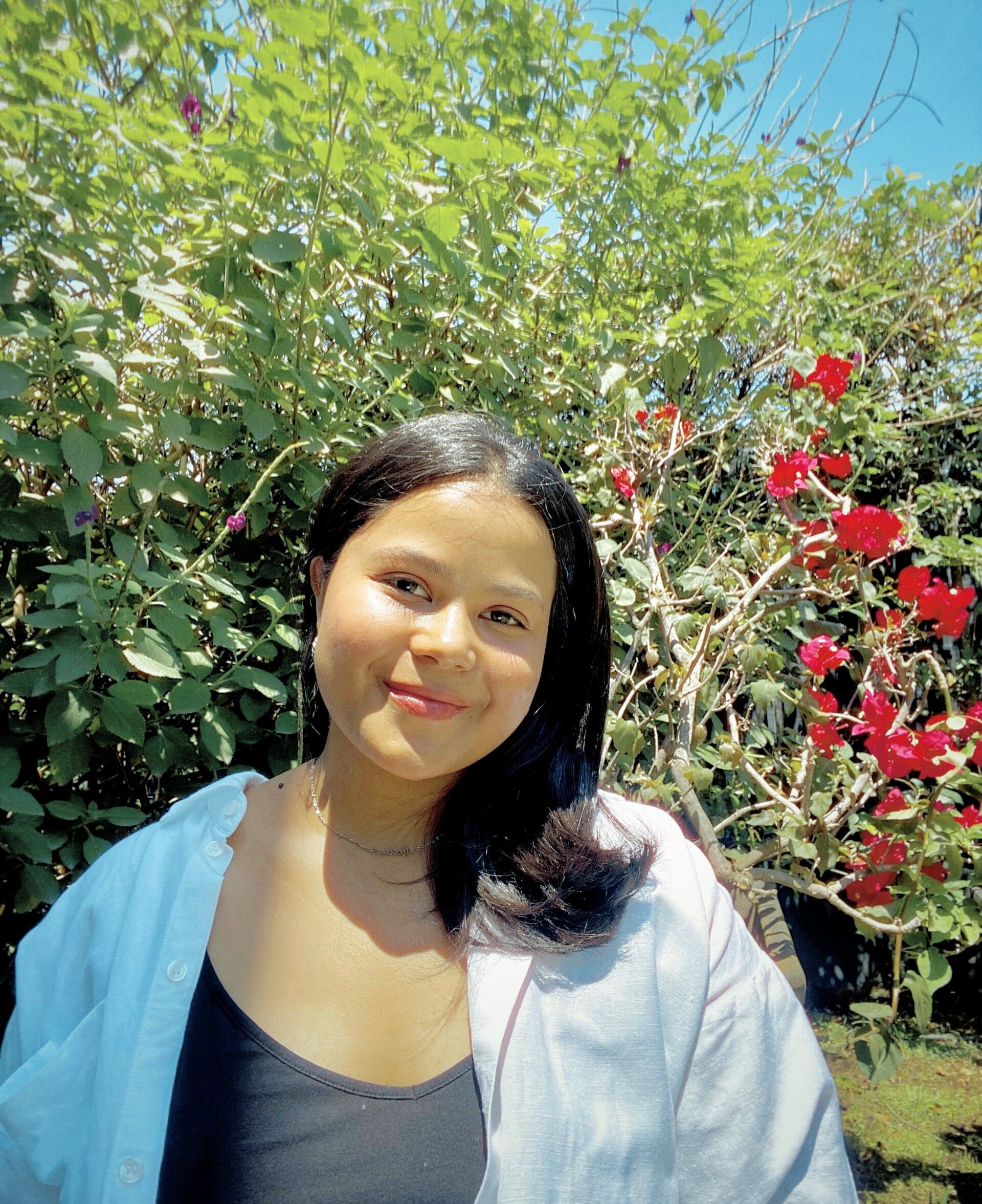 A young woman with dark hair smiles at the camera. She is wearing a black top and a white shirt. Behind her, there is lush green foliage and red flowers, suggesting an outdoor setting.
